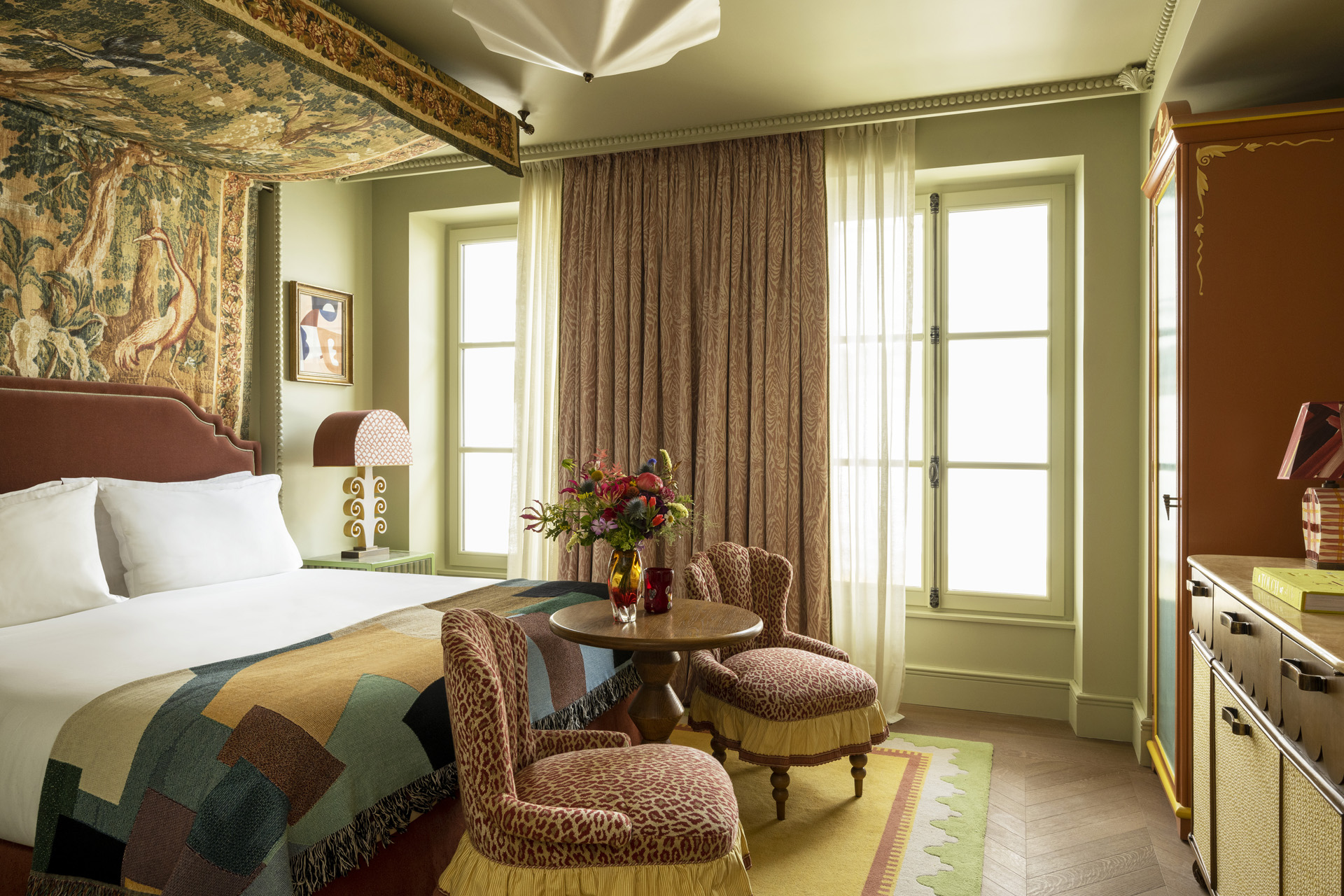 an eclectic bedroom with large bed, two sitting chairs, and two large windows